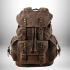 Sac à dos en toile marron foncé avec garnitures en cuir. Le sac à dos a des poches latérales et une poche frontale. Il a également des sangles en cuir et des boucles en laiton. Le fond du sac à dos est en cuir plus foncé.
— vue 8