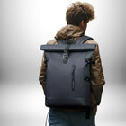 Un homme vu de dos porte un sac à dos homme imperméable gris foncé. Il porte une veste marron et un jean bleu. Le fond est flou blanc avec un dégradé.
— vue 4