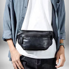 Un homme portant un sac banane cuir homme noir sur un tee-shirt blanc. Il porte une chemise bleu clair enfilée par-dessus et un jean noir. Il pose ses mains sur ses hanches.
— vue 3