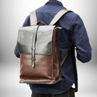 Vue arrière d'un homme portant un sac à dos urbain homme,sac à dos urbain en cuir marron et noir avec une fermeture à boucle et des bretelles. L'homme porte une chemise bleue. L'arrière-plan est un dégradé de gris. — vue 2