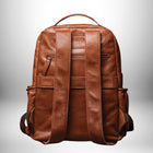 Un sac à dos en cuir marron est affiché contre un fond blanc dégradé. Le sac à dos présente des bretelles et une poignée de transport en cuir, avec plusieurs poches et fermetures éclair. La vue est oblique à l'arrière du sac à dos. — vue 3