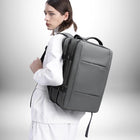 Une personne portant un sac à dos gris. La personne porte une chemise blanche et a les cheveux bruns. Le sac à dos a une étiquette "BANGE" en bas à droite. — vue 5