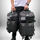 L'image montre une personne portant deux **sac a dos camping** de camouflage. Les sacs à dos ont des patchs du drapeau américain et des sangles sur le devant. La personne porte une chemise blanche et un pantalon noir.
— vue 2