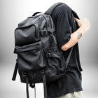 Une personne porte un sac à dos ergonomique noir. Le sac à dos est grand avec de nombreuses poches et sangles. La personne porte un t-shirt noir et a un tatouage sur le bras. — vue 2