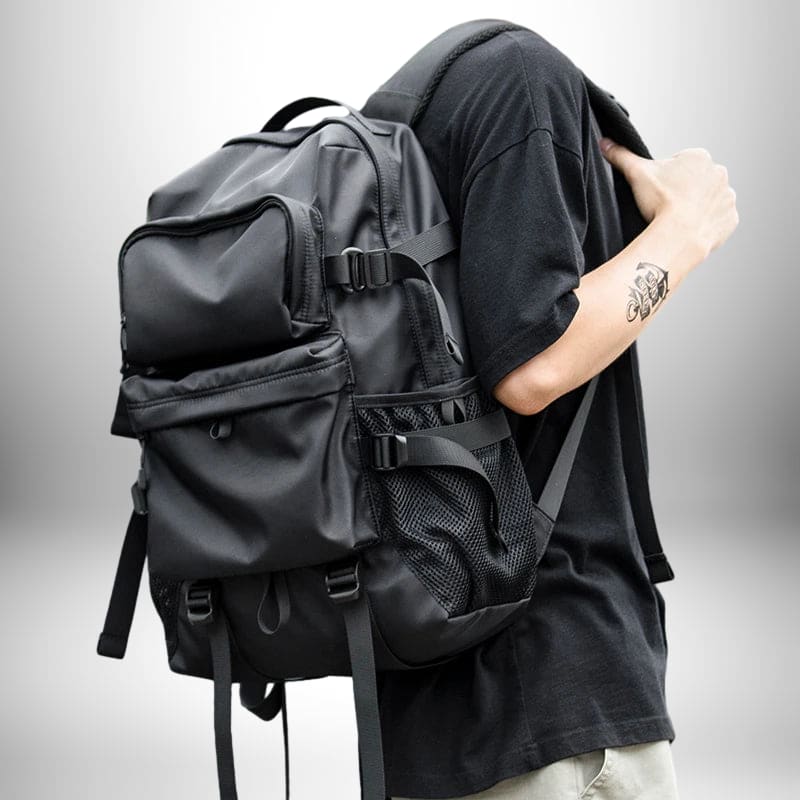 Une personne porte un sac à dos ergonomique noir. Le sac à dos est grand avec de nombreuses poches et sangles. La personne porte un t-shirt noir et a un tatouage sur le bras.