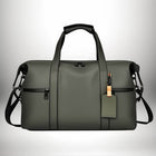 A dark green duffel bag with black accents is shown against a gradient white background. It has two handles on top, a removable shoulder strap, and a small luggage tag. — vue 5