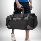 Un homme tient un grand sac sport noir et gris, apparemment en déplacement. Ses mains sont enroulées autour des poignées du sac, tandis que la partie inférieure du corps, y compris les jambes et les pieds en baskets, est visible.
— vue 6