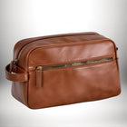 Une trousse de toilette de voyage en cuir marron, avec une fermeture éclair et une poignée latérale, posée sur une surface blanche. — vue 1