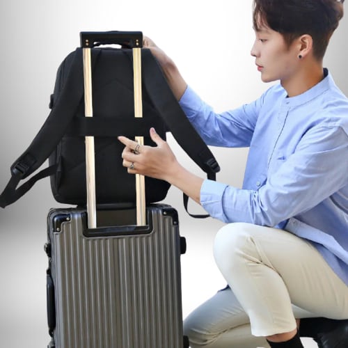 Un homme porte un sac à dos noir sur le dessus d'une valise grise. Il est assis en position accroupie, avec un bras s'étendant pour ajuster la position du sac à dos. L'homme porte un chemisier bleu et un pantalon blanc.
