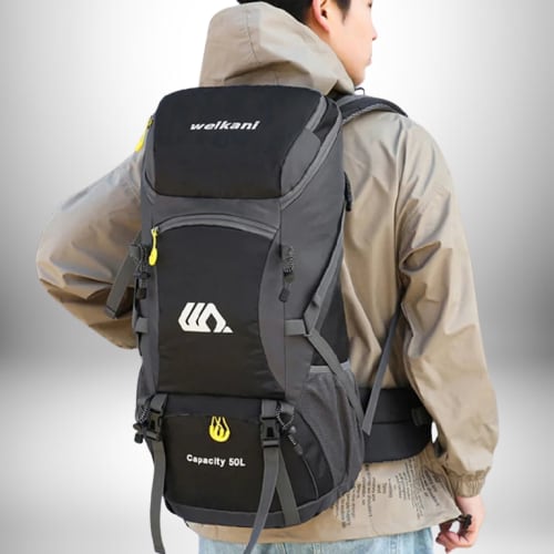 Un homme porte un sac à dos de randonnée noir et gris. Le sac à dos est marqué « welkani » en haut. Il possède également un logo blanc et jaune en haut et des poches latérales en filet gris. Le sac à dos indique « Capacite 50 L ». L'homme porte une veste beige à capuche et un jean.