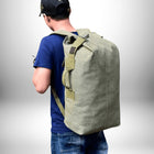 Une personne portant un grand sac à dos en toile de couleur olive. La personne est vue de dos, portant une chemise bleue, un jean et une casquette de baseball noire.
 — vue 3