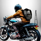 Un motard, vu de derrière, porte un casque noir et une veste en cuir marron sur une moto noire et argentée. Il roule avec un sac à dos moto rigide doté d'yeux orange sur le dos. — vue 4