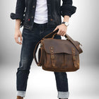 Un homme pose avec un sac en cuir style "cartable prof". Il porte une veste en jean, un tee-shirt blanc et un jean foncé retroussé. Il tient le sac par la poignée supérieure.
 — vue 3
