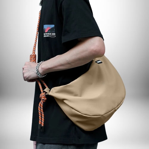 Un homme porte un petit sac de sport homme beige qui contraste avec son t-shirt noir. Le sac a une sangle en corde orange et blanche. Il porte également un bracelet en argent.
