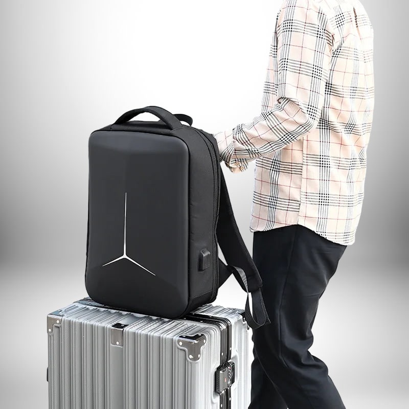 Un homme tient un sac à dos coque rigide noir, posé sur une valise argentée. Le sac à dos est orné d'un motif blanc géométrique. L'homme porte une chemise à carreaux et un pantalon noir.