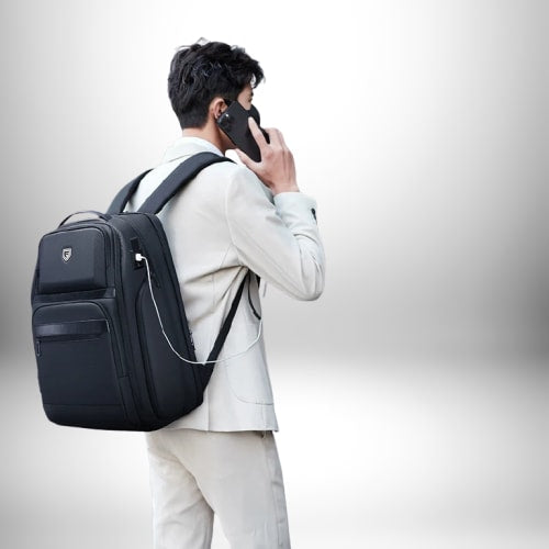 Image d'un homme, dos à la caméra, portant un sac à dos professionnel homme tout en parlant au téléphone. Il porte un costume clair et le sac à dos noir est équipé d'un port USB.
