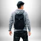 Une personne porte un sac à dos porte skate noir avec une capuche grise et un pantalon camouflage. La personne est vue de dos. — vue 3