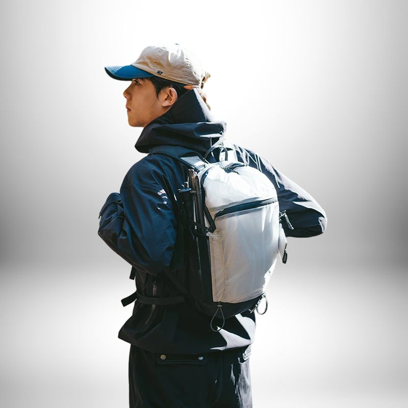 Un homme regarde par-dessus son épaule, portant un sac à dos trail blanc et gris et une casquette de baseball. Il porte également une veste noire et un capuchon.