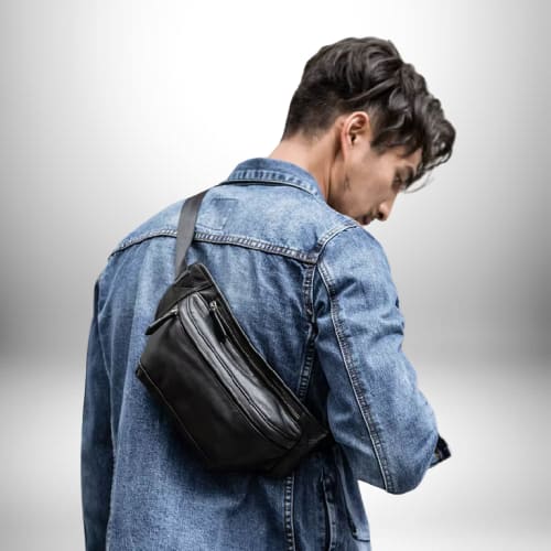 Un homme porte un sac banane cuir homme​ noir sur le dos, par-dessus une veste en jean. Le fond est blanc avec un léger dégradé de couleurs.
