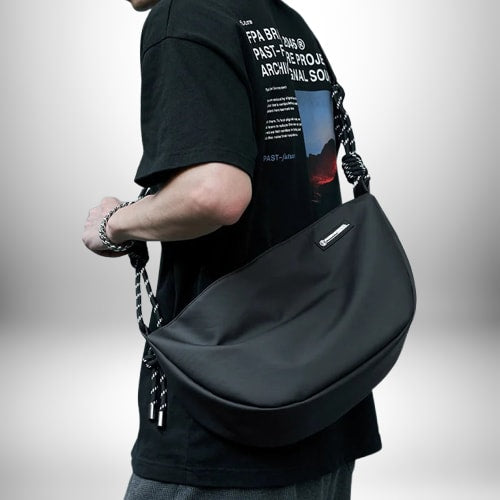 Un homme porte un petit sac de sport homme noir, avec une bandoulière torsadée noire et blanche. Il porte également un bracelet en argent et un t-shirt noir avec du texte blanc.