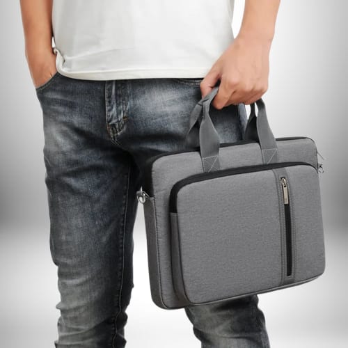 Un homme porte une sacoche ordinateur homme tendance grise devant lui. Il porte un jean délavé et un t-shirt blanc.
