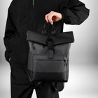 Une personne porte un sac a dos business noir, de style business, sur un fond blanc. Le sac à dos a une poche avant zippée, une boucle de fermeture centrale et une poignée supérieure. — vue 2