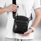 Gros plan d'un homme tenant un sac bandoulière cuir noir homme​ qui est noir. Il tient un téléphone blanc à l'intérieur du sac. Il porte une montre-bracelet blanche et un t-shirt blanc. — vue 3