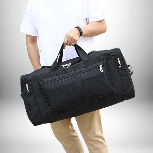 Un homme tenant à deux mains un grand sac homme sport noir. Il porte un t-shirt blanc et un pantalon kaki. 
