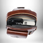 Vue en plongée à l'intérieur d'une sacoche en cuir marron, contenant un téléphone noir, un porte-cartes de crédit noir, une batterie externe blanche et un petit livre gris. Le sac a une poche à l'avant. L'arrière-plan est un gradateur gris et blanc. — vue 4