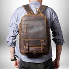 Une personne vue de dos porte un sac à dos en cuir marron foncé. Le sac à dos a une grande poche zippée à l'avant, deux petites poches avec des boucles et des sangles en dessous, ainsi que des poches latérales. La personne porte une chemise bleue à manches longues et un bracelet sur le poignet gauche. Le fond est dégradé, du blanc au gris. — vue 3
