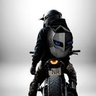 Une personne porte un sac à dos moto rigide noir et argent tout en étant assis sur une moto. Le sac à dos a des "yeux" bleus éclairés. La personne porte un casque noir et regarde vers l'avant. — vue 2