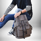 Une personne assise portant un t-shirt noir, un t-shirt à rayures blanches et noires et un jean tient un sac à dos couleur taupe avec des sangles marron, le tout sur fond dégradé blanc. La personne porte aussi une montre à bracelet. On aperçoit une des chaussures Converse Chuck Taylor All-Stars de la personne. — vue 4