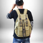 Une personne portant un sac à dos beige de style rétro. La personne porte un chapeau de baseball et un t-shirt noir avec des manches rayées blanches et noires. Elle porte également une montre-bracelet et un jean. L'arrière-plan est un simple dégradé de gris à blanc.
 — vue 3