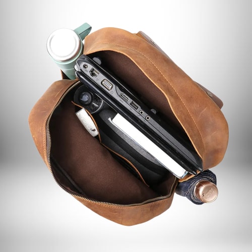 Image d'un sac à dos camel​ en cuir, ouvert, avec un ordinateur portable, une bouteille thermos, un carnet, des stylos et un parapluie plié à l'intérieur.