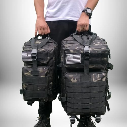 L'image montre une personne portant deux **sac a dos camping** de camouflage. Les sacs à dos ont des patchs du drapeau américain et des sangles sur le devant. La personne porte une chemise blanche et un pantalon noir.

