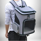 Un homme partiellement visible porte un sac à dos de transport pour animaux de compagnie gris clair. Le sac possède plusieurs compartiments, dont un compartiment principal avec une fermeture éclair et des panneaux en filet pour la ventilation. C'est un sac a dos transport chat. Sur le côté du sac, il y a une petite poche zippée. L'homme porte un t-shirt blanc à manches longues et un pantalon noir.
 — vue 5