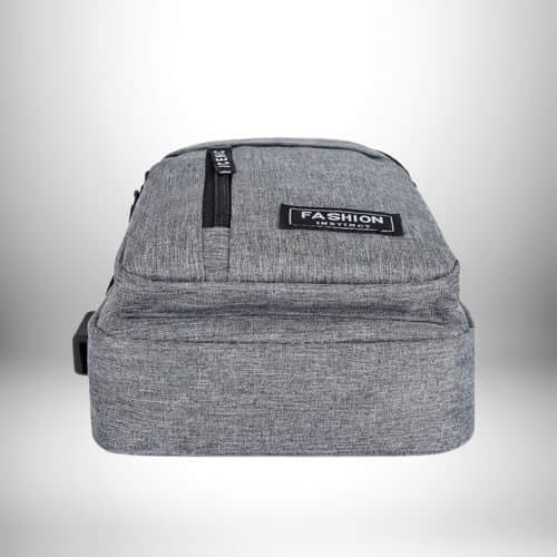 Un sac de sport bandoulière gris clair, posé sur une surface blanche et lumineuse. Le sac est de forme rectangulaire, avec une poche zippée verticale sur le devant et l'inscription "FASHION INSTINCT" sur un patch rectangulaire.
