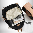 Vue de dessus d'un sac à dos en coton noir partiellement ouvert, avec en contenu un parapluie replié, une tablette, un téléphone et quelques livres. Un haut-parleur en bois est partiellement visible sur le côté droit, sur un journal avec le mot "MODE" imprimé.
 — vue 6
