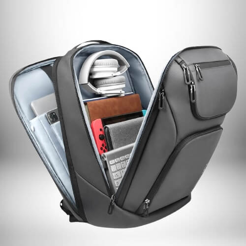 Un sac à dos professionnel homme gris sur fond blanc est montré ouvert, révélant son contenu : des écouteurs sur l'intérieur, un ordinateur portable replié, des carnets, une console, un clavier et d'autres accessoires. Le sac à dos a des poches zippées sur le côté.