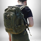 L'image montre un sac à dos randonnée homme vert foncé. Il est porté par une personne, vue de dos, portant un t-shirt noir et une casquette de baseball assortie. Le sac à dos est volumineux, avec de nombreuses poches, sangles et boucles.
 — vue 7