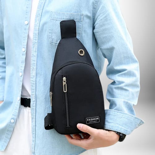 Un homme blanc tient un sac banane bandouliere homme noir. Il porte une chemise en jean boutonnée et une chemise blanche. Il porte également une montre noire à son poignet.
