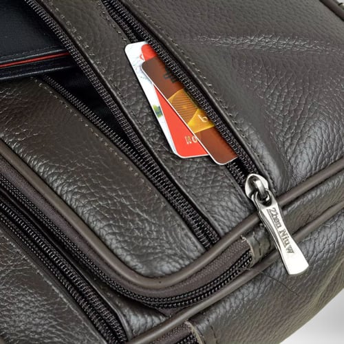 Gros plan sur un sac bandoulière homme cuir souple marron foncé avec plusieurs compartiments à fermeture éclair. Des cartes de crédit sont à moitié insérées dans une poche ouverte. La tirette en métal doré d'une des poches est gravée des lettres « Zhen Niw ». Le cuir est texturé.
