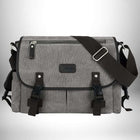 Un sac bandoulière en toile homme de couleur grise avec des sangles et des boucles noires est disposé sur un fond gris clair. Le sac a une poche zippée sur le dessus et a une conception à rabat. Il a également des boucles sur le devant pour le fixer.
 — vue 8