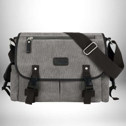 Un sac bandoulière en toile homme de couleur grise avec des sangles et des boucles noires est disposé sur un fond gris clair. Le sac a une poche zippée sur le dessus et a une conception à rabat. Il a également des boucles sur le devant pour le fixer.
