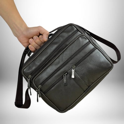 L'image montre un sac bandoulière homme cuir souple​ marron foncé avec plusieurs poches zippées. Une main tient la poignée du sac. Le fond est un dégradé de gris clair.

