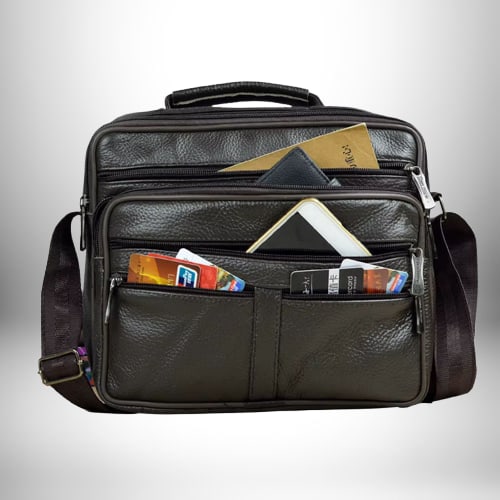 Un sac bandoulière homme cuir souple​ brun foncé avec plusieurs poches zippées à l'avant et sur le dessus, contenant un portefeuille noir, un téléphone, des cartes de crédit et des documents. Une bandoulière en tissu marron foncé est attachée.

