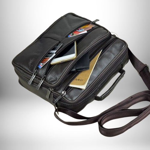 Un sac bandoulière homme cuir souple marron, avec de nombreuses poches zippées et contenant divers objets tels qu'un téléphone, des cartes de crédit, un portefeuille et un carnet. Le sac est équipé d'une bandoulière réglable.
