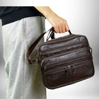 Un homme tient un sac bandoulière homme cuir souple. Le sac est de couleur marron foncé avec plusieurs poches zippées. L'homme porte des vêtements gris et un pull noir qui ne couvrent qu'en partie le corps du modèle. Le fond est dégradé de clair à blanc.
 — vue 7