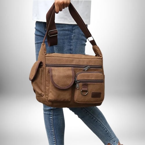 Une personne porte à la main un sac de ville homme en toile marron, avec plusieurs poches et une bandoulière.

