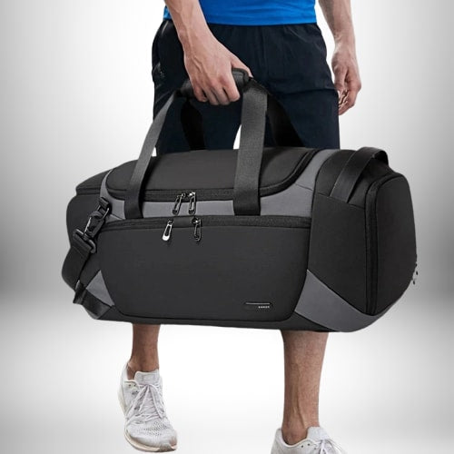 Un homme tient un grand sac sport noir et gris, apparemment en déplacement. Ses mains sont enroulées autour des poignées du sac, tandis que la partie inférieure du corps, y compris les jambes et les pieds en baskets, est visible.  
